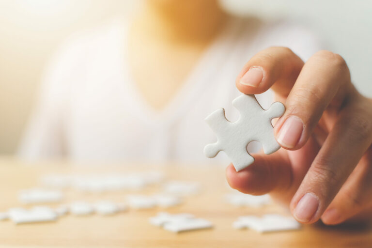 Hand of female trying to connect pieces of white jigsaw puzzle on wooden table.